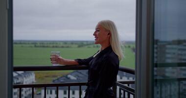 girl stands on her balcony engaged in a phone conversation using headphones while sipping water from a glass. She enjoys the view of beautiful green fields and an urban landscape video