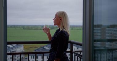 Young girl stands on a balcony talking on her phone through earbuds while sipping water. The view showcases green fields and modern buildings in the background on a cloudy day video