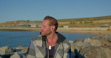 athletic man with a beard stands on the seashore talking on his phone while wearing headphones. The sun is shining and the beach is visible in the background creating a peaceful atmosphere video