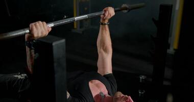 man is focused on lifting a heavy barbell during bench press exercises. He is wearing headphones and is training in a well equipped gym environment video
