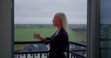 Girl stands on a balcony wearing headphones while talking on the phone. She holds a glass of water and enjoys the calm view of the fields and distant houses video