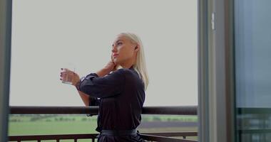 girl stands relaxed on a balcony holding a glass of water while conversing on her phone with headphones on. The scene captures a tranquil atmosphere amidst an open view video
