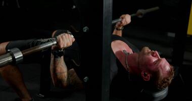 In a gym during the evening a man is engaged in a bench press workout while wearing headphones. He demonstrates concentration and effort as he lifts the barbell video