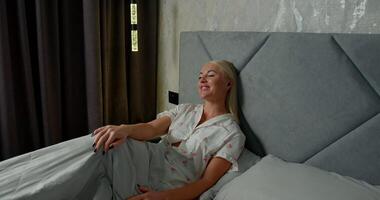 girl sits comfortably in bed wearing headphones and smiling while talking on the phone. Sunlight gently filters through the curtains creating a cozy atmosphere at home video