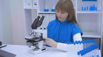 A focused lab technician in a blue uniform and gloves prepares scientific samples in a modern laboratory. The image includes a microscope in the foreground, test tubes, and a clean, organized video