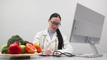 Shot of beautiful smart nutritionist woman working with computer while taking notes in the nutritionist consultation. Concept of healthy eating, vegetarianism and weight loss video