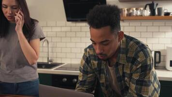 Multicultural couple in the kitchen during a quiet morning. He focuses on his laptop while she speaks on the phone. A salad in progress lies on the table as they share a calm routine. video
