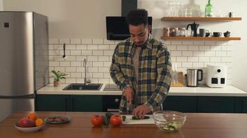 African-American man calmly preparing a salad in the kitchen during a quiet morning. He slices cucumber, adds it to a bowl, and tastes a piece with a soft smile. video