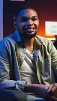 Vertical Portrait of african american person smiling and working in his living room, using laptop to solve remote freelancing tasks as a self employed guy. Young adult feels relaxed at home. Camera A. video