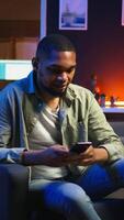 Vertical African american person texting messages on a smartphone app on the sofa, finishing tasks before relaxing at home. Young adult scrolling his mobile phone, chatting via texts. Camera A. video