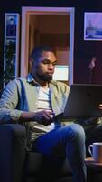 Vertical African american person attending a webinar on wireless pc in his living room, relaxing at home with freelance work. Guy engaging in a discussion, multitasking to finish daily tasks. Camera A. video