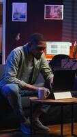 Vertical Adult guy typing on laptop laying on couch in stylish apartment, engaging in freelancing and online work. Black person multitasking efficiently and embracing flexibility of remote job. Camera A. video