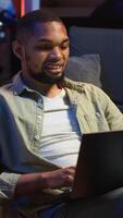 Vertical Adult guy typing on laptop laying on couch at his stylish loft space, engaging in freelancing and online work. Black person multitasking and embracing the flexibility of remote job. Camera B. video
