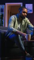 Vertical Self employed guy browsing the web and listening to music, having fun with songs on headset and completing tasks. African american man laying on sofa and relaxing in the living room. Camera A. video
