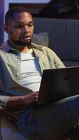 Vertical Skilled african american man multitasking on laptop in cool living room, connected to the internet while managing freelance work and remote responsibilities. Browsing web on bean bag. Camera B. video