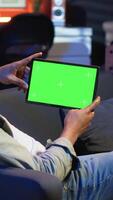 Vertical African american male freelancer holding phone with mockup copy space screen, relaxing on sofa in a space with neon lights and smart tv setup. Young person feels casual at home. Camera B. video