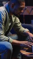 Vertical Adult guy typing on laptop laying on couch in stylish apartment, engaging in freelancing and online work. Black person multitasking efficiently and embracing flexibility of remote job. Camera B. video