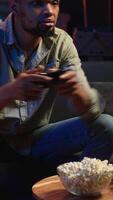 Vertical Male gamer eating popcorn while he plays online shooting tournament, having fun with games in his free time at home. Relaxed guy enjoying game night with snacks and competition. Camera B. video