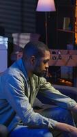 Vertical African american freelancer listening to a podcast being focused on online work, using headphones while seated on the couch in his living room. Multitasking in a casual home. Camera A. video