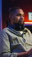 Vertical Black person listening to focus music on headphones to finish tasks, multitasking in his living room on the bean bag. Young adult enjoying calm study sounds to complete assignments. Camera A. video