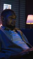Vertical Skilled african american man multitasking on laptop in cool living room, connected to the internet while managing freelance work and remote responsibilities. Browsing web on bean bag. Camera A. video