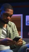 Vertical Relaxed black man scrolling on his smartphone in a comfortable space, sitting at the coffee table. Modern home highlights leisure and online connectivity in a stylish apartment. Camera A. video