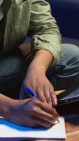 Vertical Black freelancer taking notes on textbook in order to solve his tasks, doing research and studying articles online. Young adult enjoying remote job and relaxed schedule, telework. Camera B. video