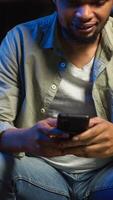 Vertical Carefree freelancer chatting with friends over text messages on phone, sitting on the couch and scrolling social media apps. Black guy showcases staying entertained while working remotely. Camera B. video