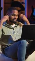 Vertical Black person listening to focus music on headphones to finish tasks, multitasking in his living room on the bean bag. Young adult enjoying calm study sounds to complete assignments. Camera B. video