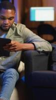 Vertical Black remote worker navigating on tablet next to screen running analytics, sitting at home working on his daily tasks with multiple devices. Leisure and job productivity in loft. Camera B. video