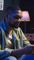 Vertical Carefree freelancer chatting with friends over text messages on phone, sitting on the couch and scrolling social media apps. Black guy showcases staying entertained while working remotely. Camera A. video