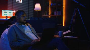 Adult man typing on laptop while he lays on a bean bag at home, engaging in freelancing and online work. Black guy multitasking efficiently and embracing the flexibility of remote job. Camera A. video