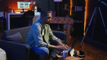Focused African American guy teleworking on his laptop in a stylish living room. Remote worker balancing comfort and work efficiency in a modern workspace. Comfortable loft studio. Camera A. video