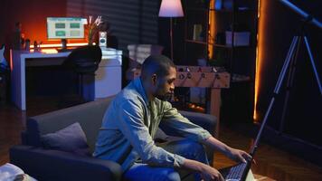 African american freelancer listening to a podcast being focused on online work, using headphones while seated on the couch in his living room. Multitasking in a casual home. Camera A. video
