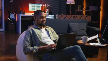 Adult guy typing on laptop laying on couch at his stylish loft space, engaging in freelancing and online work. Black person multitasking and embracing the flexibility of remote job. Camera A. video