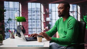 African american adult typing on a computer in his loft apartment, engaging in online freelancing work and multitasking efficiently. Flexibility of remote work in a cozy home office. Camera B. video