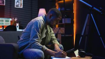 Black freelancer taking notes on textbook in order to solve his tasks, doing research and studying articles online. Young adult enjoying remote job and relaxed schedule, telework. Camera A. video