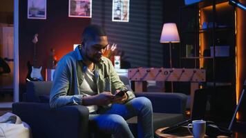 Smiling young adult chatting with friends over texts on mobile phone, sitting on the couch and scrolling social media apps. Young guy staying entertained while working remotely. Camera A. video