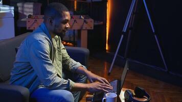 Young black adult working on his freelance tasks in the living room, adjusting his job schedule and enjoying work from his cozy home. Person navigating on web with laptop, multitasking. Camera A. video