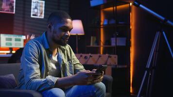 Carefree freelancer chatting with friends over text messages on phone, sitting on the couch and scrolling social media apps. Black guy showcases staying entertained while working remotely. Camera A. video