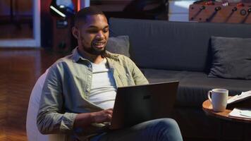 Adult guy typing on laptop laying on couch at his stylish loft space, engaging in freelancing and online work. Black person multitasking and embracing the flexibility of remote job. Camera B. video