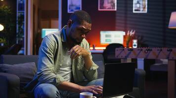 African american guy being overworked with headache at modern loft, working remotely under stress and pressure from his boss. Young person frustrated and worried, struggling with tasks. Camera A. video