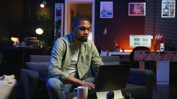 Young black guy working on his freelance tasks in the living room, adjusting his job schedule and enjoying work from his cozy home. Person navigating on web with laptop, multitasking. Camera A. video