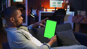 Relaxed male tech user holding device with isolated mockup screen in his living room, sitting on the couch and teleworking. African american happy guy browses the internet, outsourcing. Camera A. video