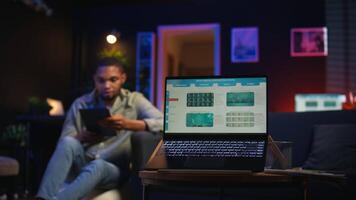 Black remote worker navigating on tablet next to screen running analytics, sitting at home working on his daily tasks with multiple devices. Leisure and job productivity in loft. Camera A. video