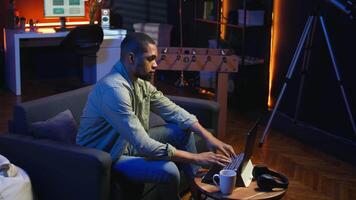 African american guy does research by reading articles and data driven papers in the living room, using online sources to collect information for a new project. Multitasking. Camera A. video