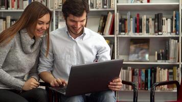 Students share a laugh while watching something on a laptop in the library video