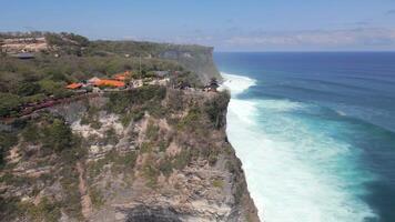 Aerial drone view of Uluwatu Temple on cliffs above the turquoise ocean in Bali, Indonesia. Famous Hindu temple and cultural landmark surrounded by crashing waves, and coastal scenery. video