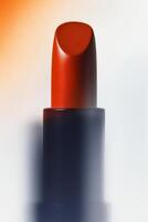 Artistic close-up of a modern orange lipstick with a rounded, smooth surface in a deep navy tube, standing vertically against a gradient background photo