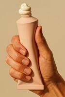 A close-up of a human hand holding a soft pastel pink cosmetic tube with a swirl of white cream on top, on a warm beige background. photo